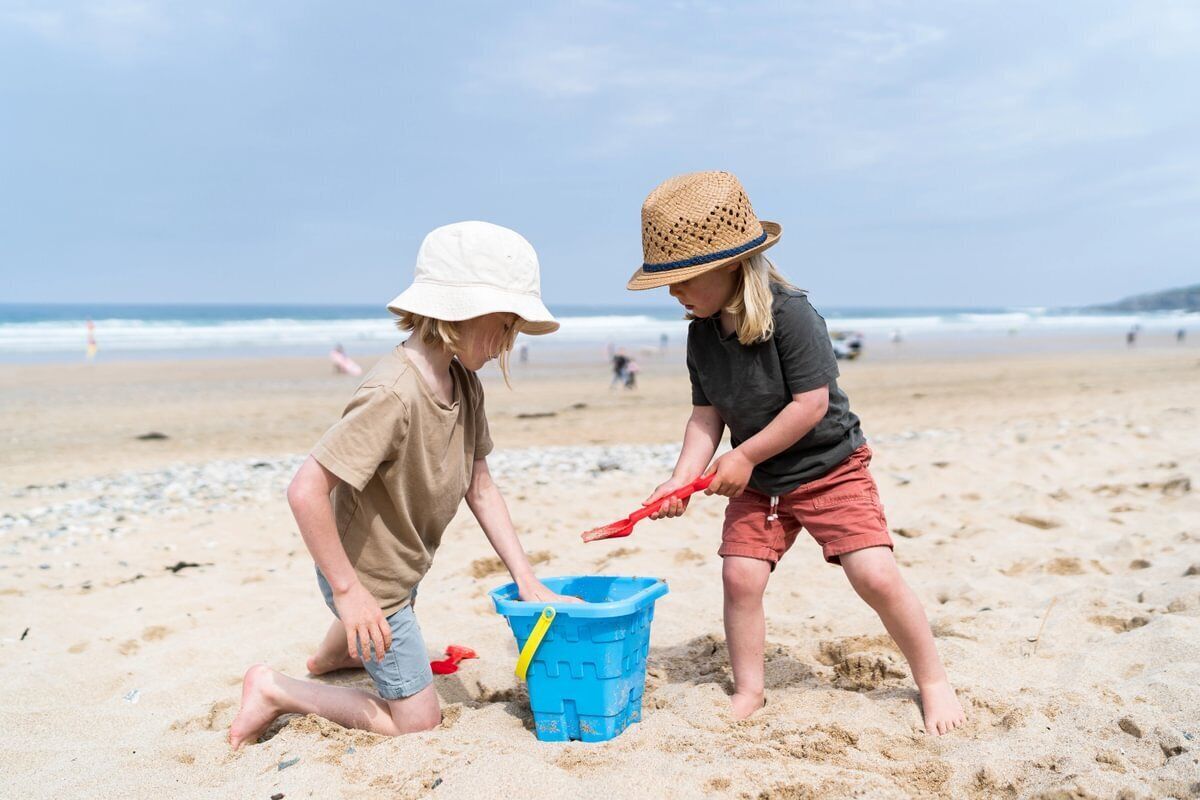 Deux enfants jouent avec un seau bleu sur une plage de sable, avec l’océan en arrière-plan.