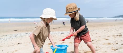 Two children playing with a blue bucket on a sandy beach with the ocean in the background.