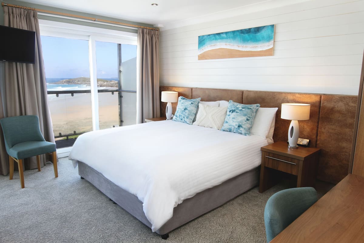 A hotel room with a large bed, two bedside tables with lamps, a chair, a TV, and a view of the ocean.