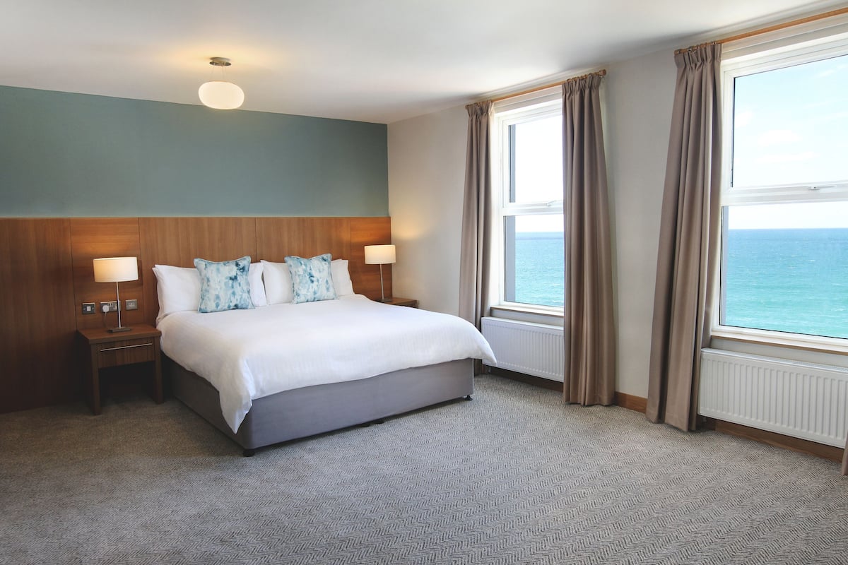 A hotel room with a large bed, two bedside tables, a nightstand, a window with curtains, and a view of the sea.
