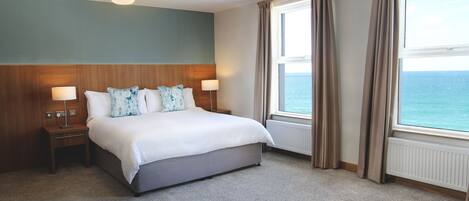 A hotel room with a large bed, two bedside tables, a nightstand, a window with curtains, and a view of the sea.