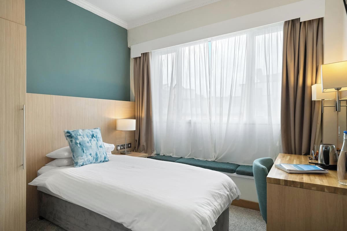 A hotel room with a bed, a desk, a chair, a window with curtains, and a lamp.