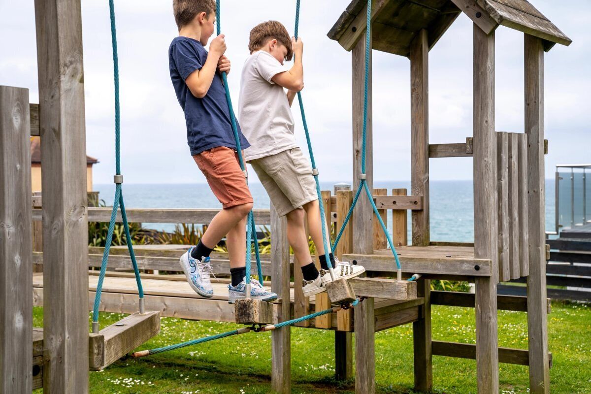 Two children on a wooden bridge with ropes, overlooking a body of water.