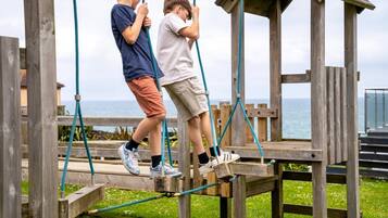 Two children on a wooden bridge with ropes, overlooking a body of water.
