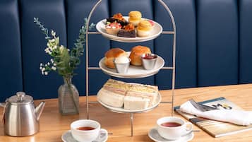 A three-tiered dessert stand with various pastries and sandwiches, a vase with flowers, a teapot, cups of tea, and a magazine on a table.