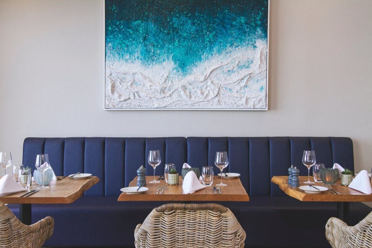 A restaurant interior with a dark blue booth seating, wooden tables with set dining ware, and a large abstract painting on the wall.