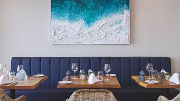 A restaurant interior with a dark blue booth seating, wooden tables with set dining ware, and a large abstract painting on the wall.