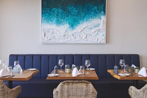 A restaurant interior with a dark blue booth seating, wooden tables with set dining ware, and a large abstract painting on the wall. - The Esplanade Hotel (Newquay)