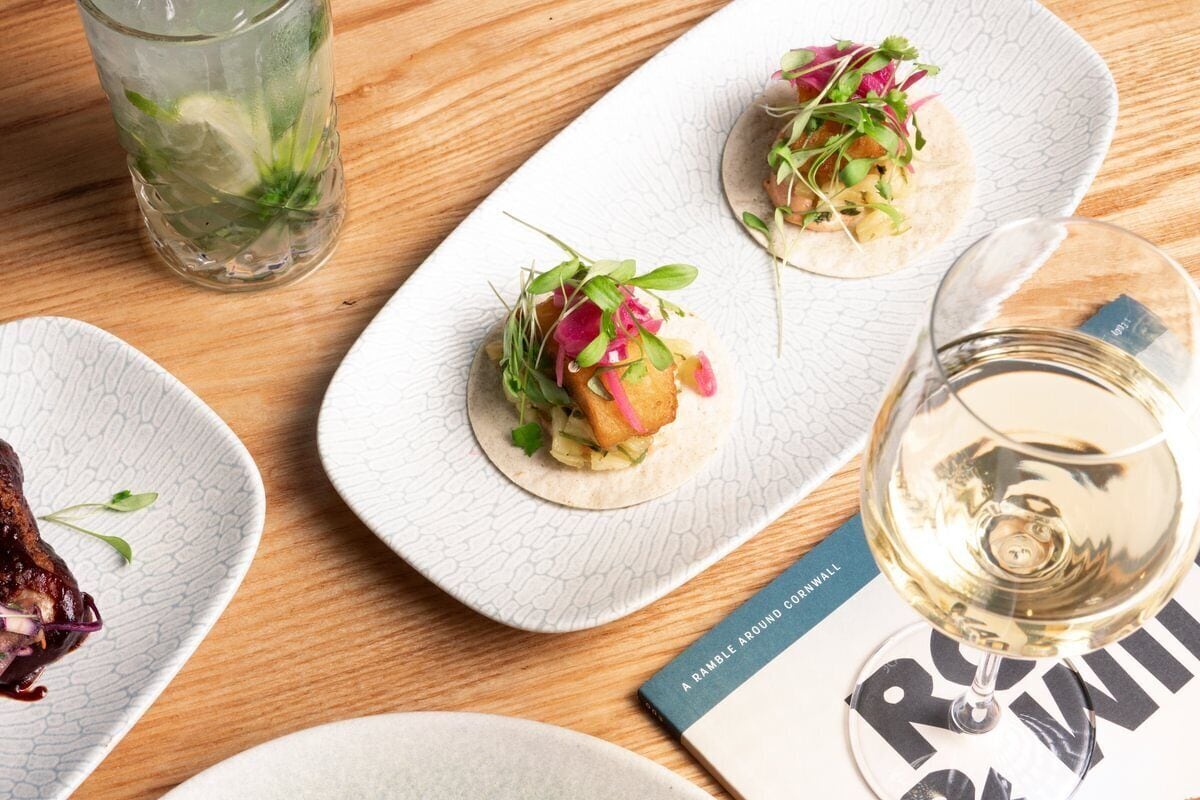 A table with plates of food, a glass of white wine, and a book titled 'A RAMBLE AROUND CORNWALL'.