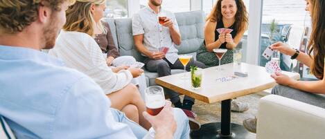 A group of people playing cards at a table with drinks, seated on a couch with a view of the sea.