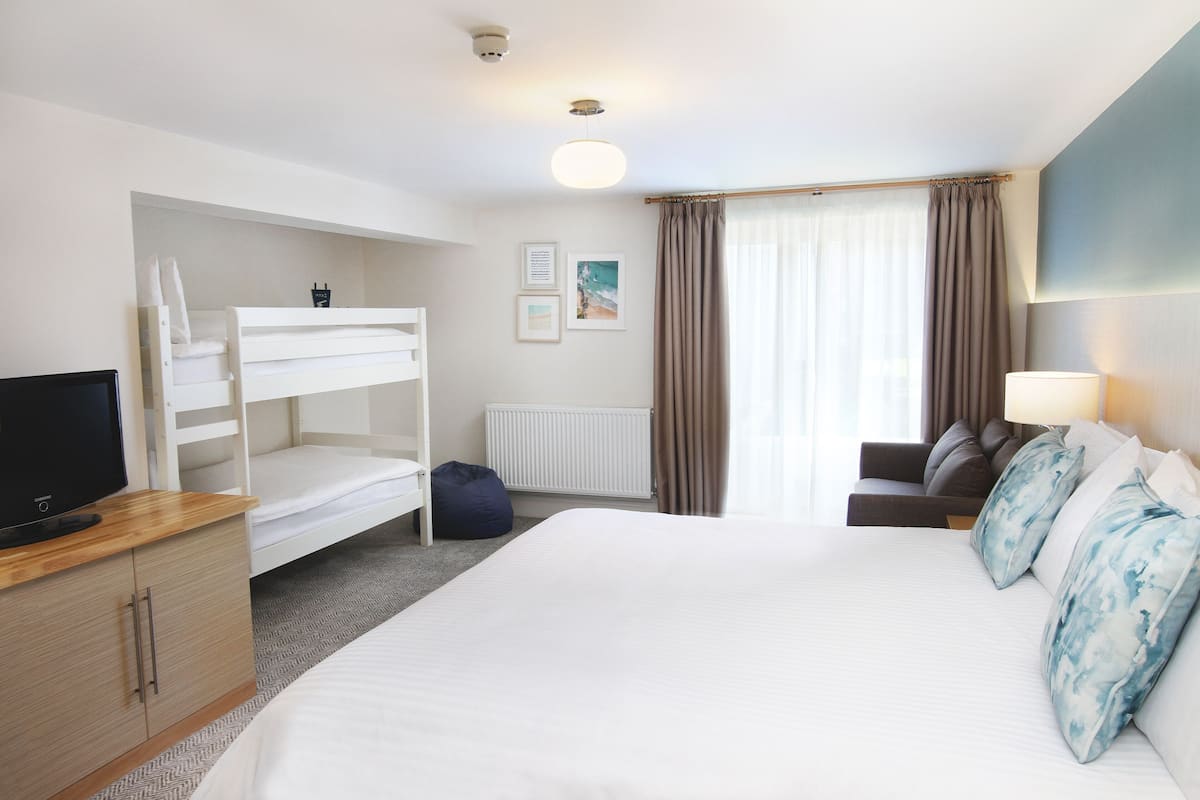A hotel room with a bunk bed, a desk with a computer, a bed with pillows, a lamp, and a window with curtains.