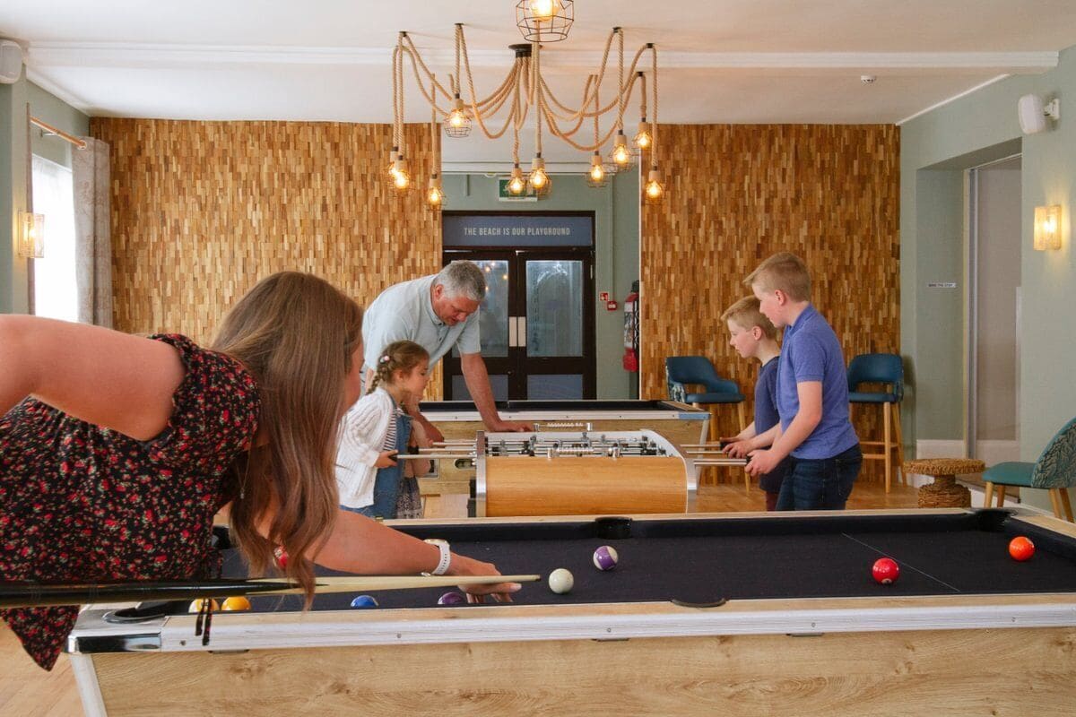 A pool table with balls, a foosball table, and a sign that reads 'THE BEACH IS OUR PLAYGROUND'.