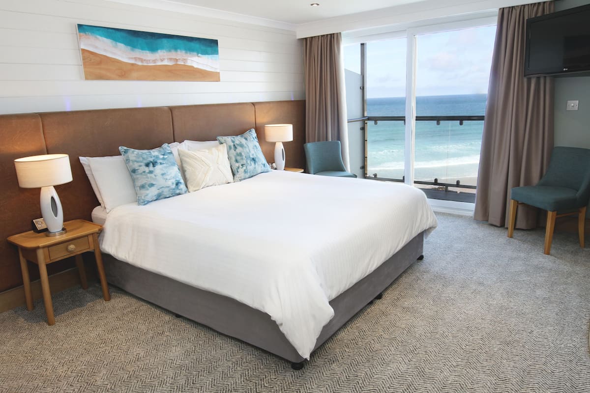 A hotel room with a large bed, two bedside tables with lamps, a chair, a TV, and a large window with a view of the ocean.