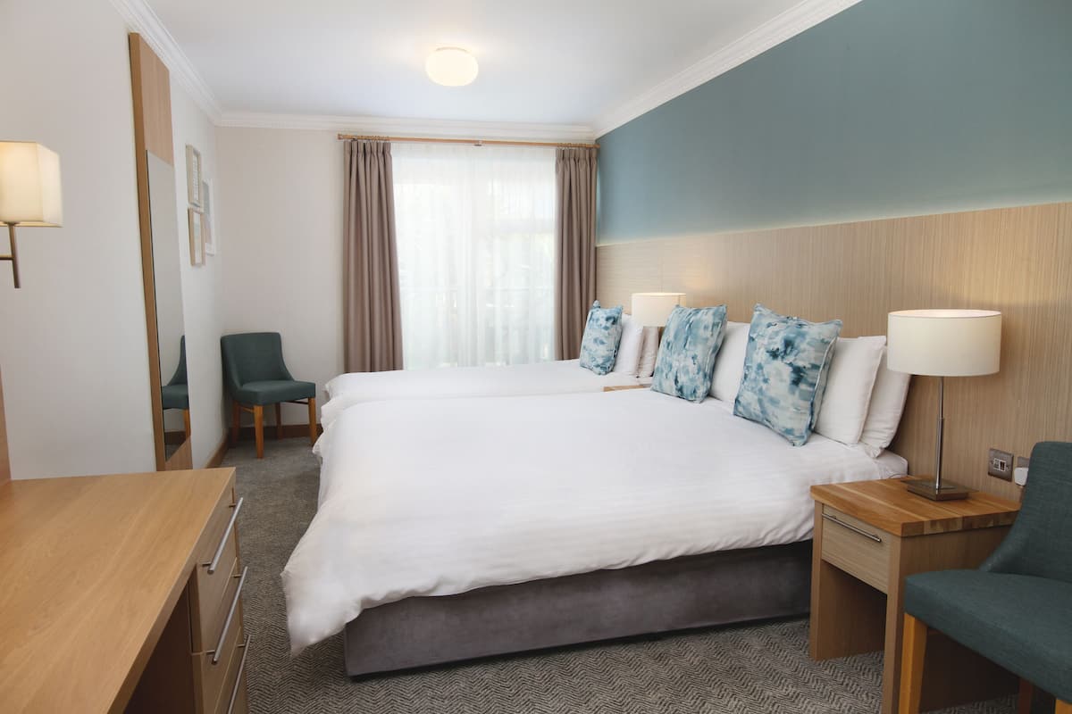 A hotel room with a large bed, two bedside tables with lamps, a desk, a chair, and a window with curtains.