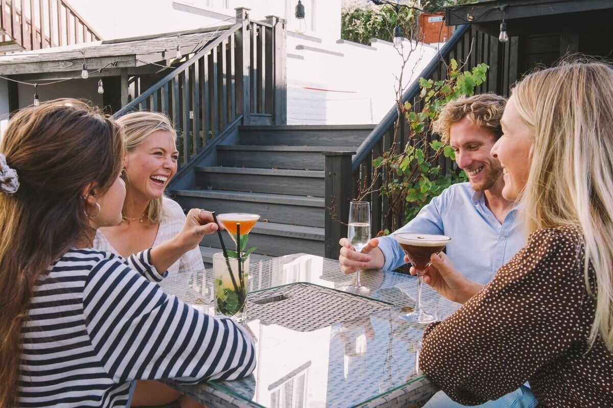 A group of people sitting at an outdoor table with drinks, a staircase in the background, and a building facade.