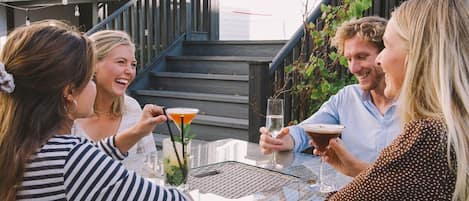 Un grupo de personas sentadas en una mesa al aire libre, con bebidas, una escalera al fondo y la fachada de un edificio.
