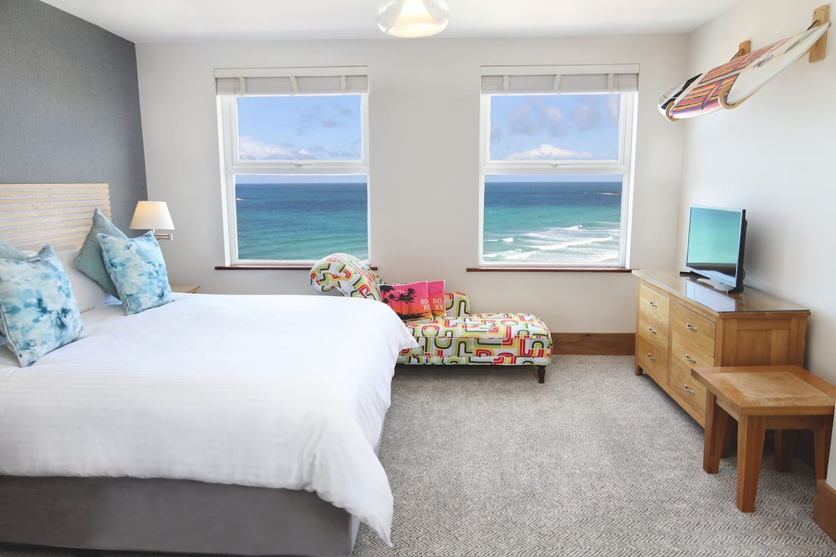 A bedroom with a bed, a desk, a chair, a TV, and a view of the ocean through two windows.