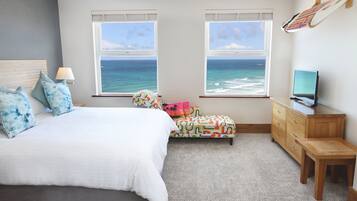 A bedroom with a bed, a desk, a chair, a TV, and a view of the ocean through two windows.