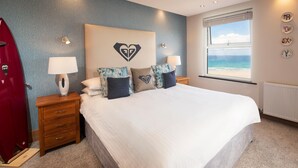 A bedroom with a bed, nightstands, a red coat, a window with a view of the beach, and a heart-shaped wall decoration. - The Esplanade Hotel (Newquay)