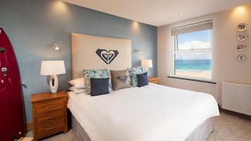 Ein Schlafzimmer mit Bett, Nachttischen, einem roten Mantel, einem Fenster mit Blick auf den Strand und einer herzförmigen Wanddekoration.