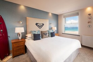 A bedroom with a bed, nightstands, a red coat, a window with a view of the beach, and a heart-shaped wall decoration. - The Esplanade Hotel (Newquay)