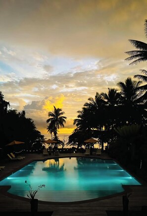 Une piscine avec vue sur le coucher de soleil, entourée de palmiers et de chaises longues.