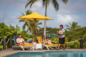 Un área junto a la piscina con sillones, una sombrilla amarilla y una persona sirviendo bebidas.