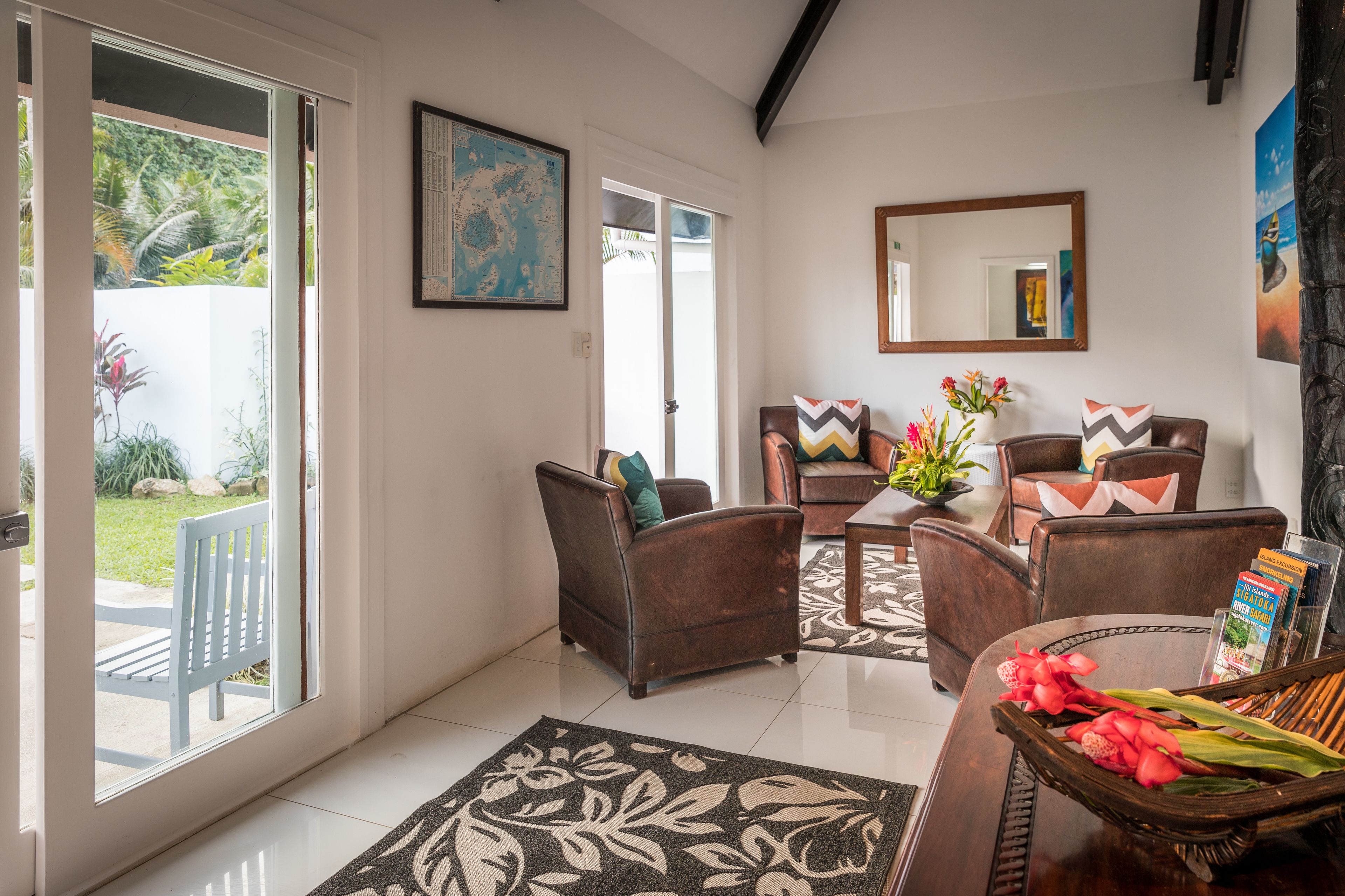 A spacious living area with a large sliding glass door leading to a garden, a comfortable seating arrangement with brown leather chairs, a coffee table, and a decorative vase with flowers.