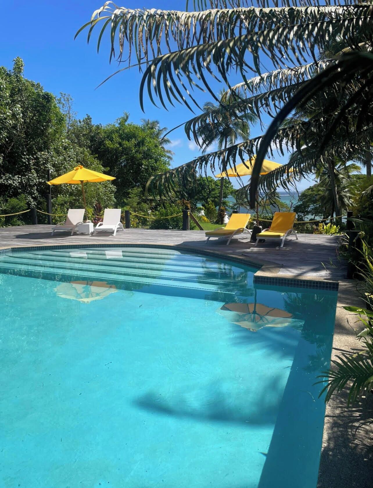 A clear blue pool with white and yellow lounge chairs and umbrellas, surrounded by lush greenery.