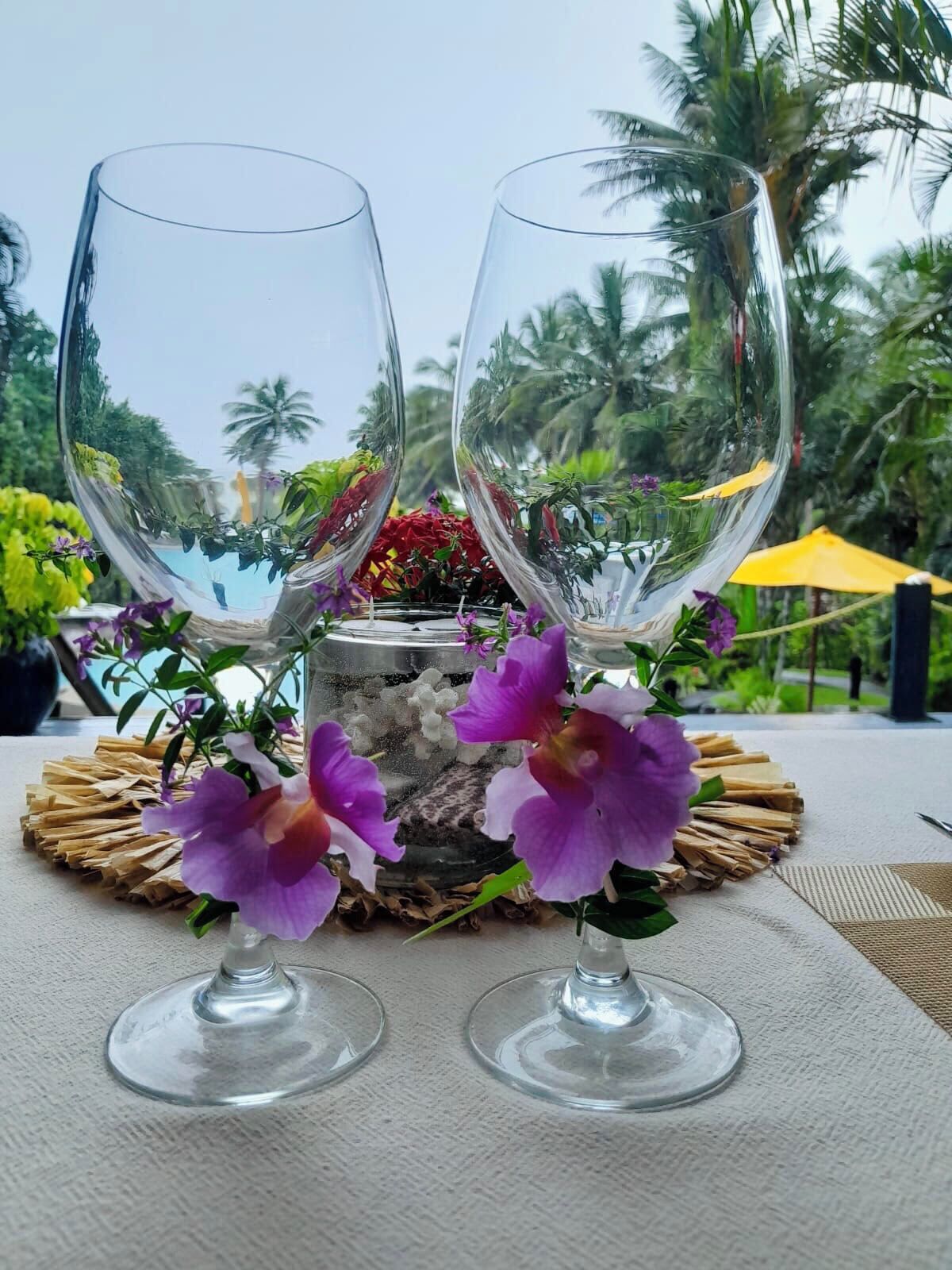 Two empty wine glasses on a table with a woven placemat, surrounded by purple flowers, with a view of palm trees and a pool in the background.