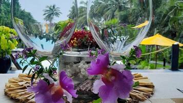 Two empty wine glasses on a table with a woven placemat, surrounded by purple flowers, with a view of palm trees and a pool in the background.