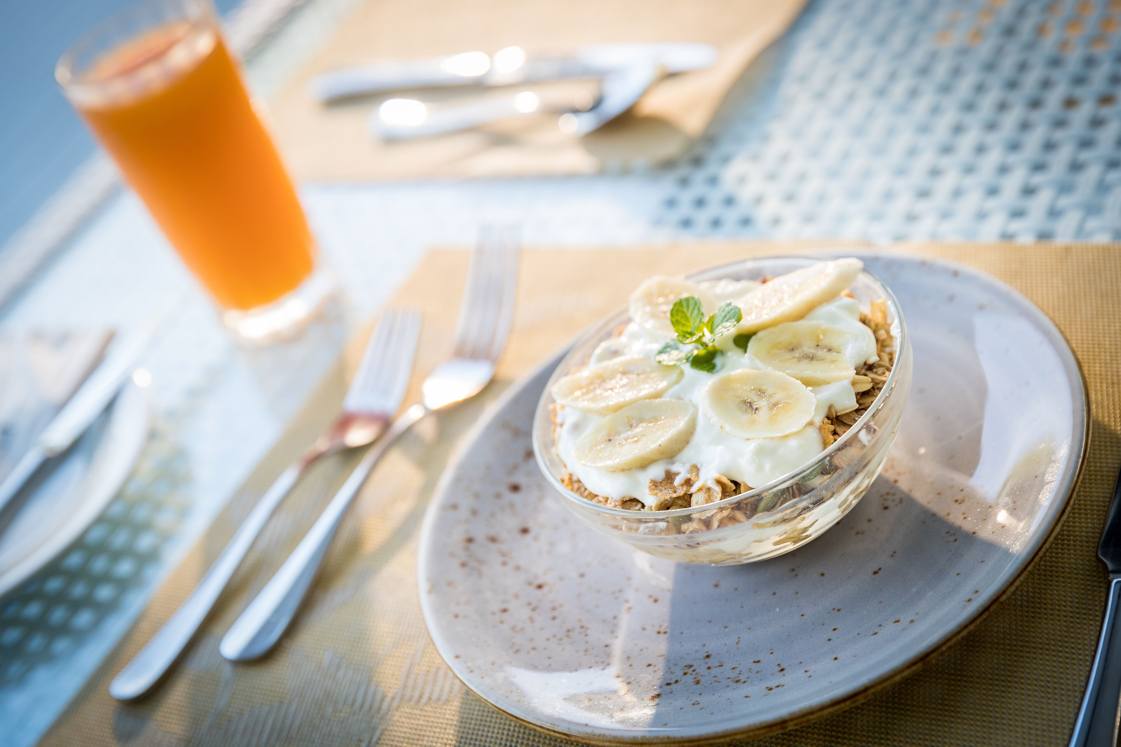 A breakfast setting with a bowl of cereal topped with banana slices and a mint leaf, a glass of orange juice, and cutlery on a table.