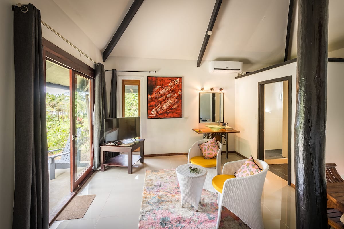 Un salon moderne avec un téléviseur, des meubles en osier blancs, un grand tableau et une vue sur la verdure à l’extérieur.
