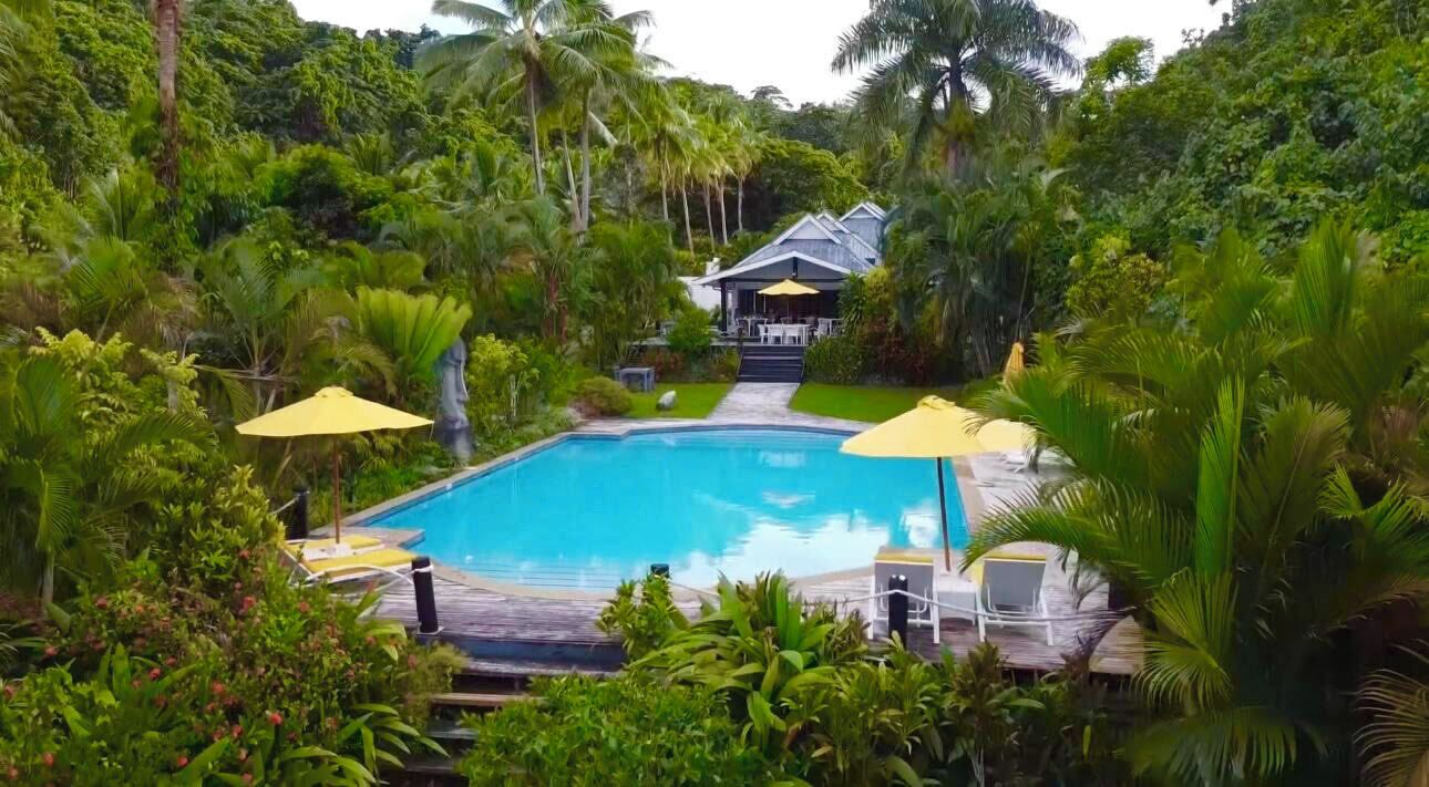A swimming pool surrounded by lush greenery, with a covered seating area and a building in the background.