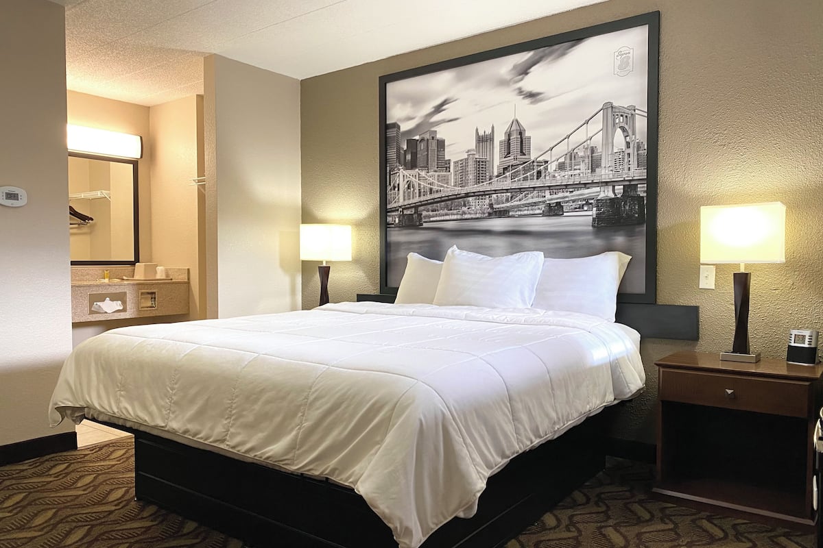 A hotel room with a large bed, two bedside tables with lamps, a nightstand, a mirror, and a cityscape painting on the wall.