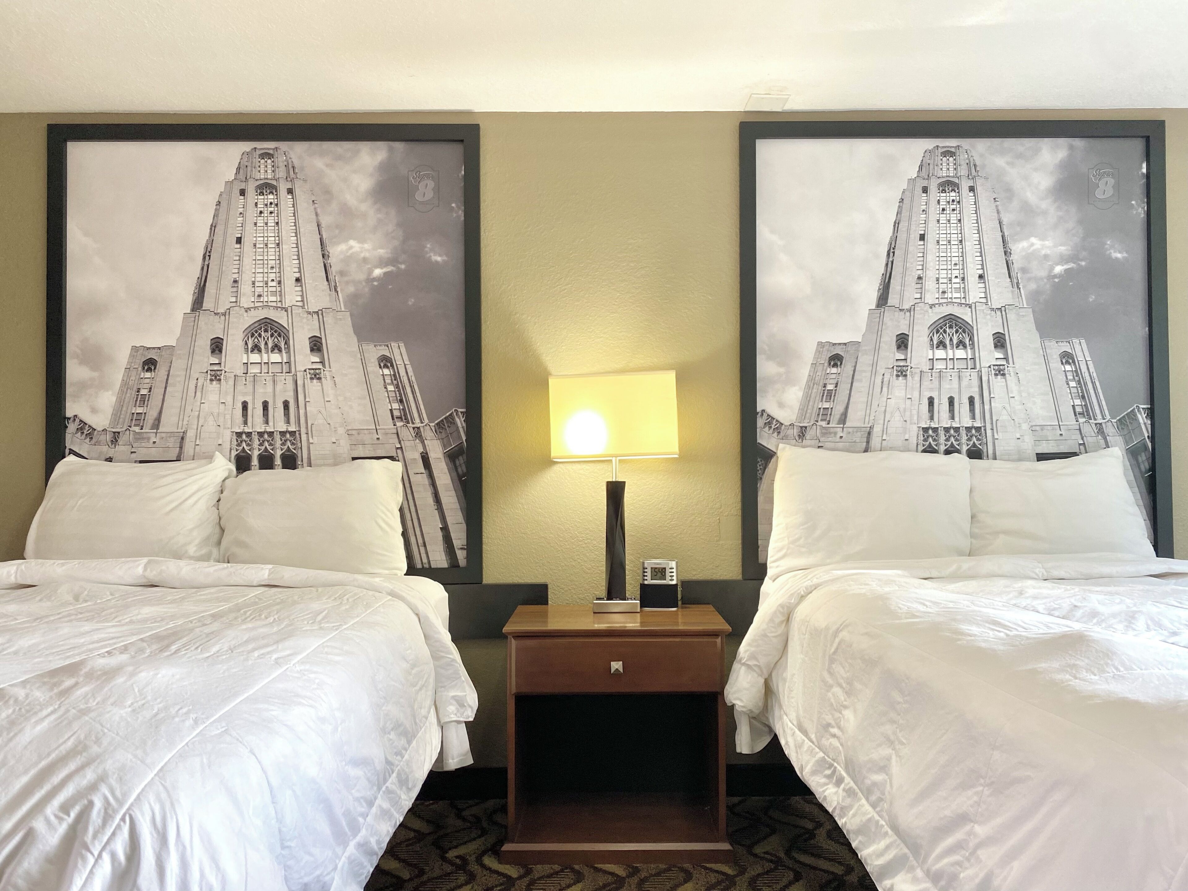 A hotel room with two beds, a nightstand with a lamp, and two large framed pictures of a building.