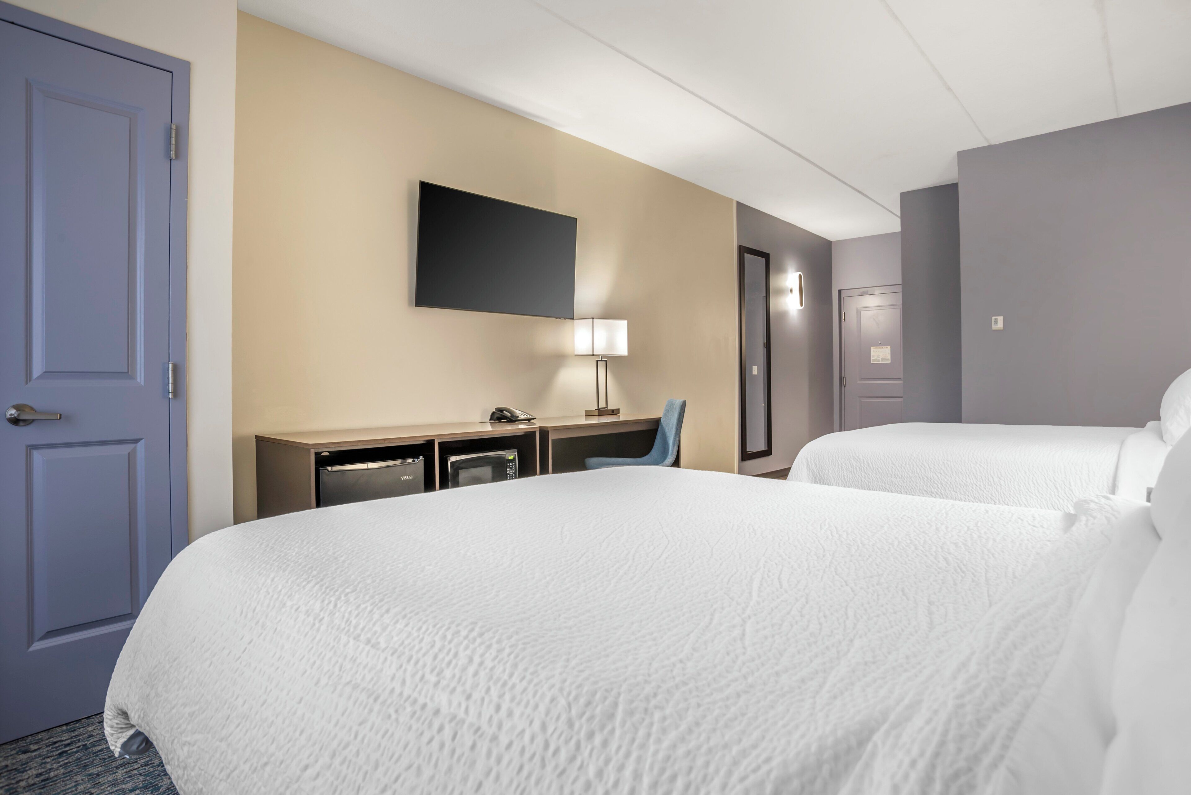 Een hotelkamer met twee bedden, een bureau met lamp, een flatscreen-tv en een minibar.