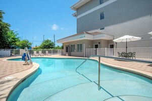 Ein Swimmingpool mit weißem Zaun, ein überdachter Bereich mit Stühlen und ein Gebäude im Hintergrund.