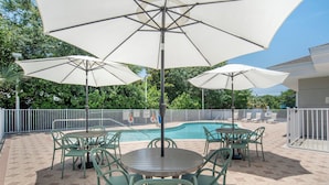 An outdoor patio with a pool, surrounded by a white fence, featuring green chairs and large umbrellas.