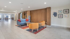 A spacious lobby with a blue sofa, orange armchairs, and a coffee table. There are wooden cabinets and a refrigerator in the background.