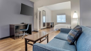 A modern hotel room with a flat-screen TV, a desk with a chair, a bed, and a sofa with a patterned pillow.