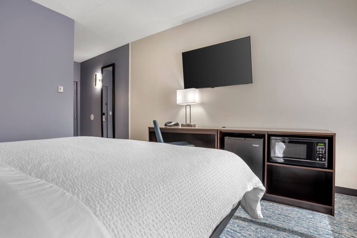 Une chambre d’hôtel équipée d’un lit, d’un bureau avec un micro-ondes, d’une lampe et d’un téléviseur à écran plat fixé au mur.