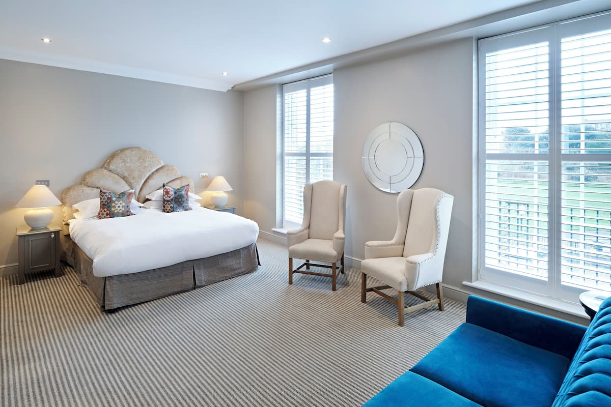 A modern bedroom with a large bed, two bedside tables with lamps, a chair, a blue sofa, and large windows with blinds.