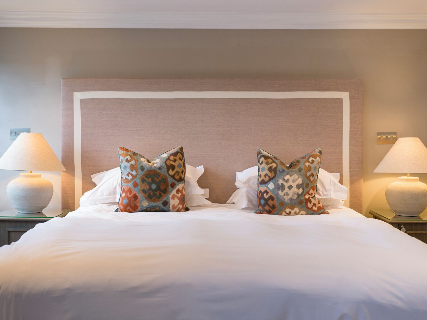 A neatly made bed with two decorative pillows, a large padded headboard, and two bedside lamps on either side.