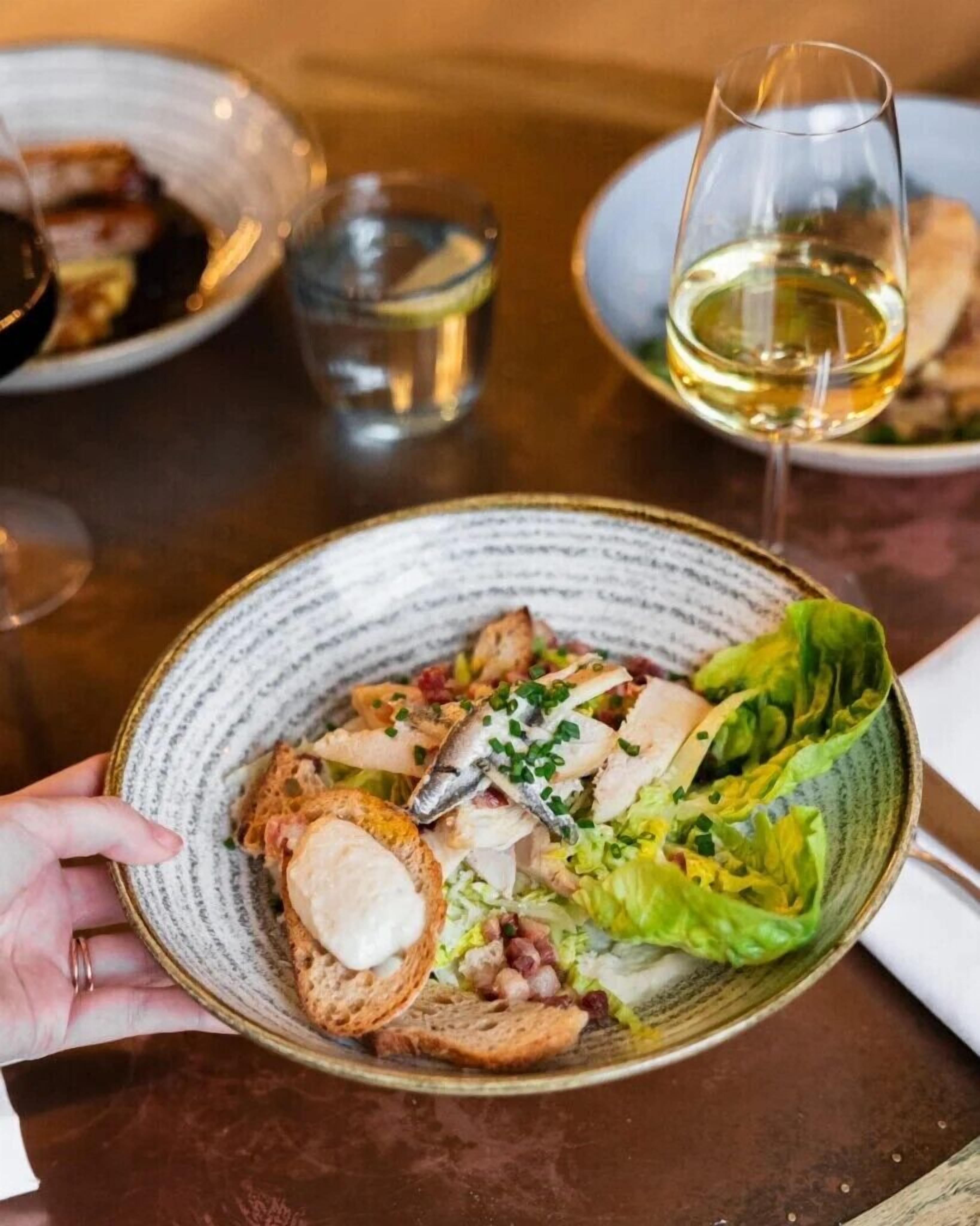A person holding a bowl of food with bread and lettuce, with a glass of white wine and a plate of food in the background.