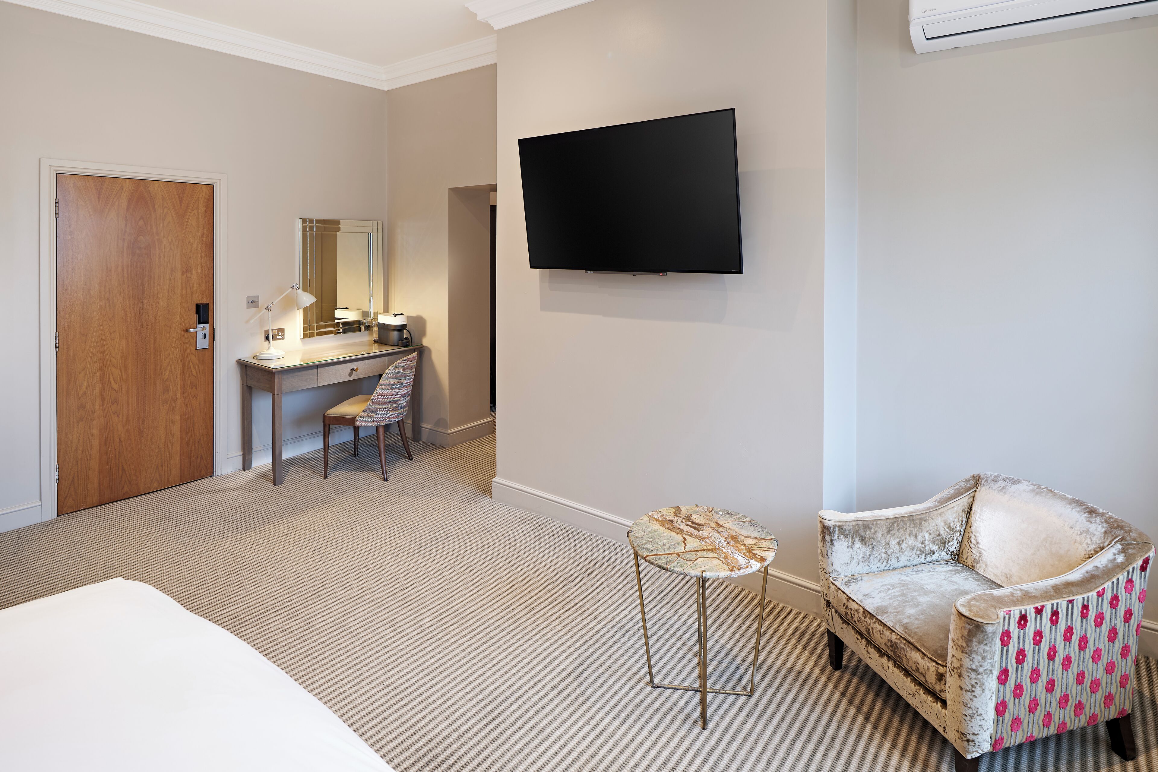 Habitación de hotel con cama, escritorio con silla, televisor montado en la pared y sillón estampado.