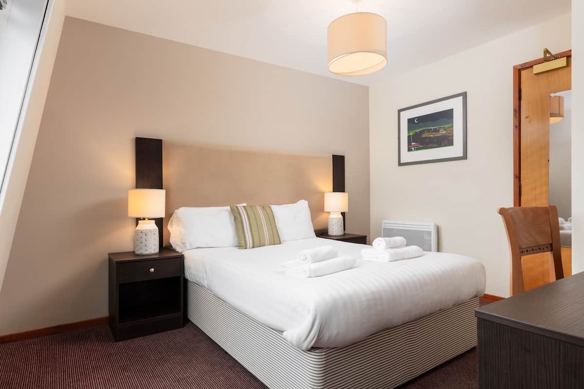 A hotel room with a large bed, two bedside tables with lamps, a wooden chair, a desk, and a framed picture on the wall.