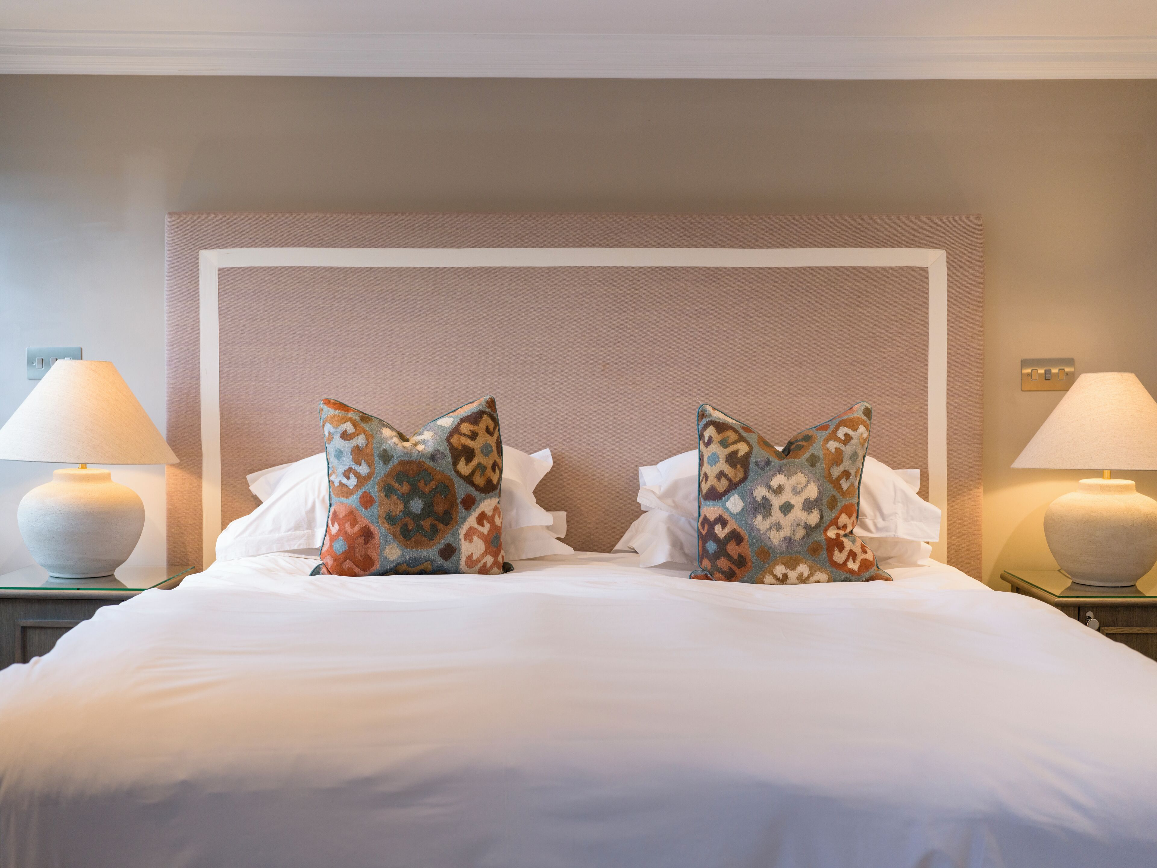 A neatly made bed with two decorative pillows, a bedside lamp, and a wall-mounted light switch.