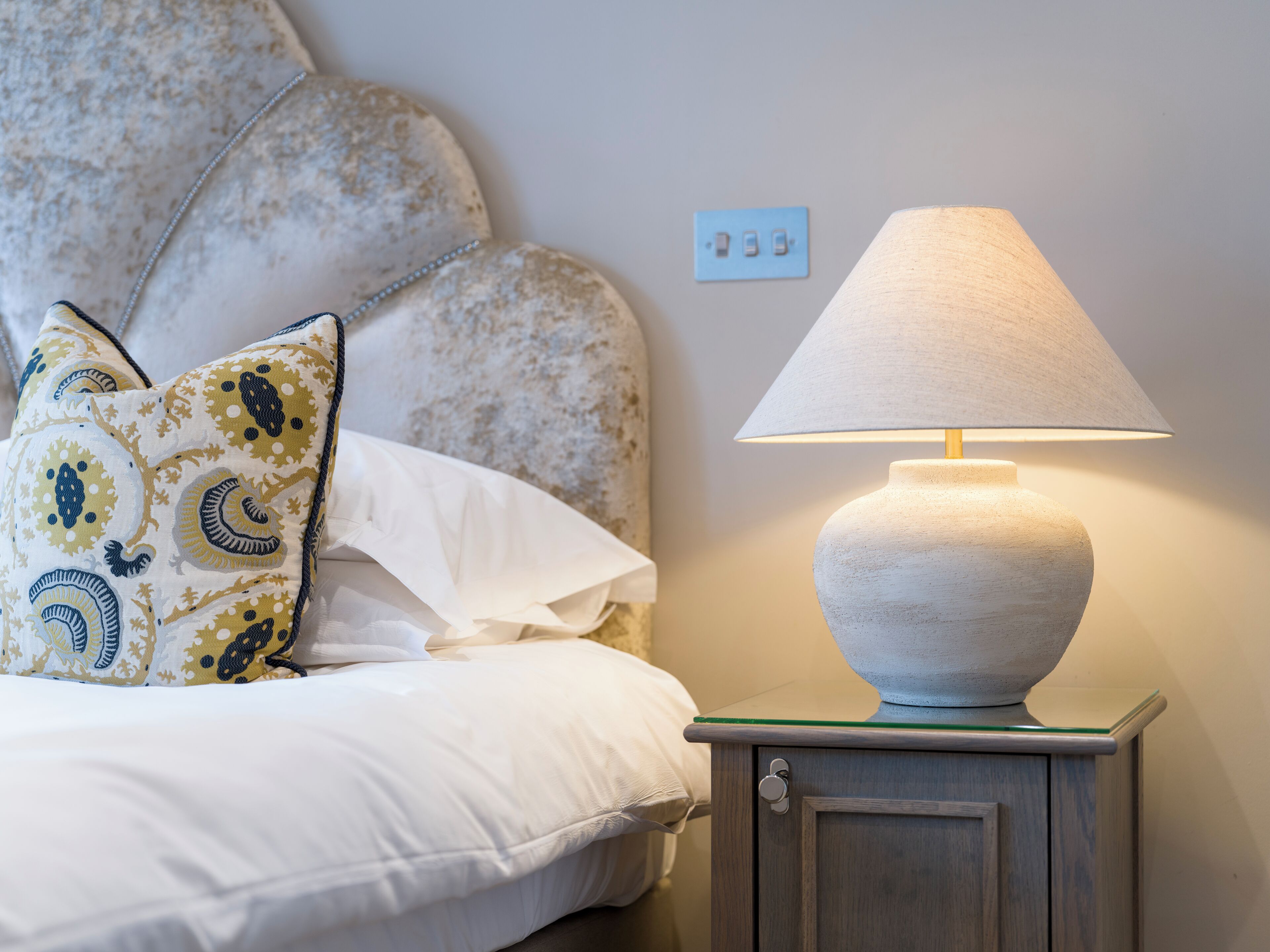 Una cama con almohadones estampados, una lámpara de noche y un enchufe de pared.