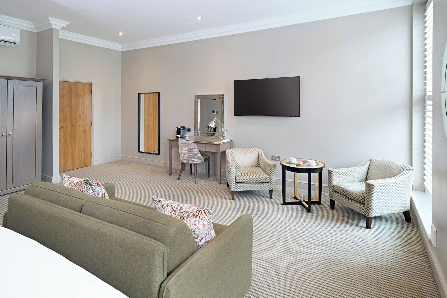 A hotel room with a sofa, two armchairs, a desk with a chair, a flat-screen TV, and a wardrobe.
